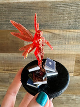 Load image into Gallery viewer, Red fairy figurine on a black stand with books against a wooden background