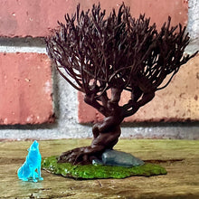 Load image into Gallery viewer, gnarled brown willow tree miniature surrounded by green grass and a grey rock. The miniature is sitting on a wood grain flat surface and the wall behind is red brick. There is a very small blue wolf miniature sitting next to the willow.